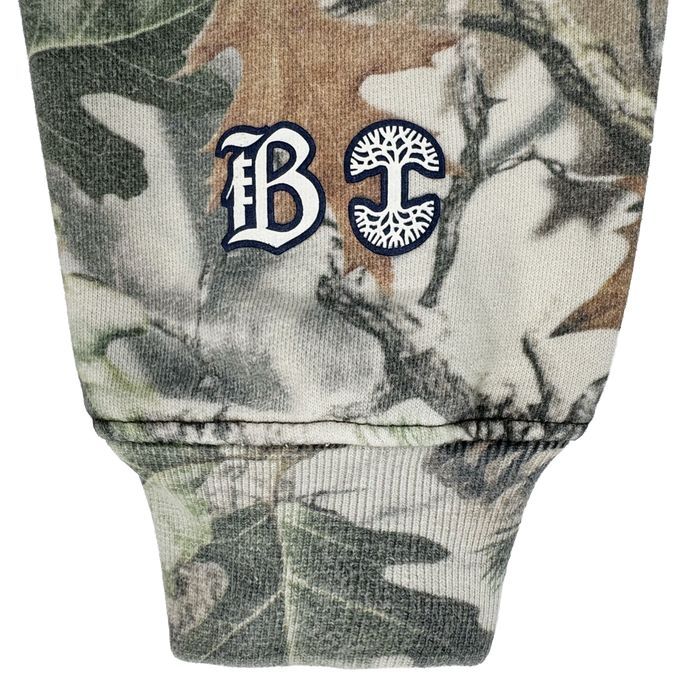 A close-up of the Bay FC Camo Hoodie shows a cuffed sleeve with green, brown, and beige camouflage. Near the seam are white and navy embroidered gothic "B" and a "C" with a tree design, representing Bay FC’s branding.