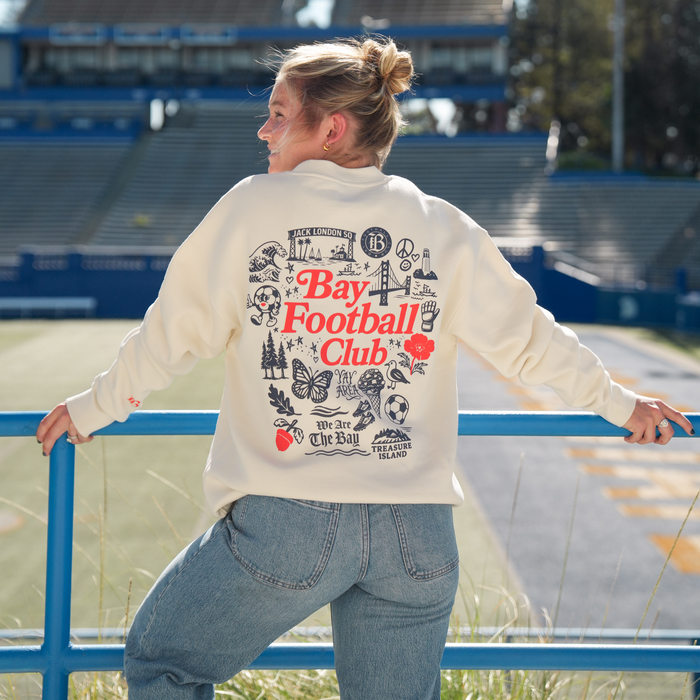 A person with blonde hair in a ponytail stands by a blue railing at an empty stadium, smiling over their shoulder. They're wearing the Bay FC Jubilee Crew sweatshirt from Bay FC and light blue jeans.