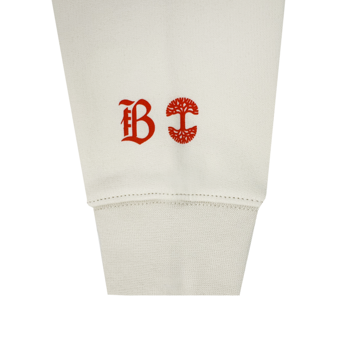 A close-up of the Bay FC Jubilee Crew shows its off-white cuff and sleeve, featuring two red symbols: Bay FC’s stylized “B” logo and a circular tree emblem, printed side by side above the cuff.