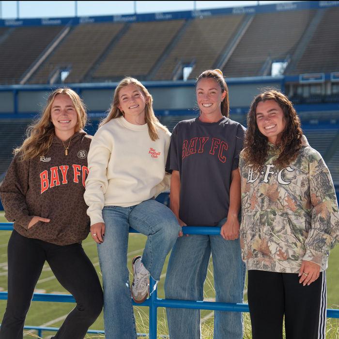 Four women stand smiling at a blue railing in a stadium. Two wear Bay FC Jubilee Crews, one wears a Bay FC T-shirt, and another sports a Bay FC camo hoodie. Empty bleachers and a green field fill the background.