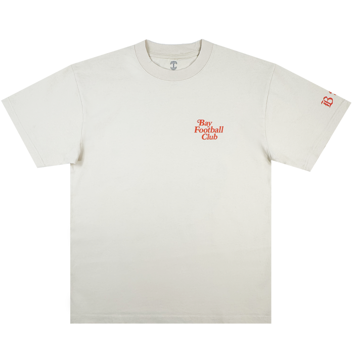 The Bay FC Jubilee Tee is a classic fit, heavy-weight cotton shirt in off-white, featuring "Bay Football Club" in small orange chest text and a matching "B" logo on the left sleeve. It has a crewneck and short sleeves. Brand: Bay FC.