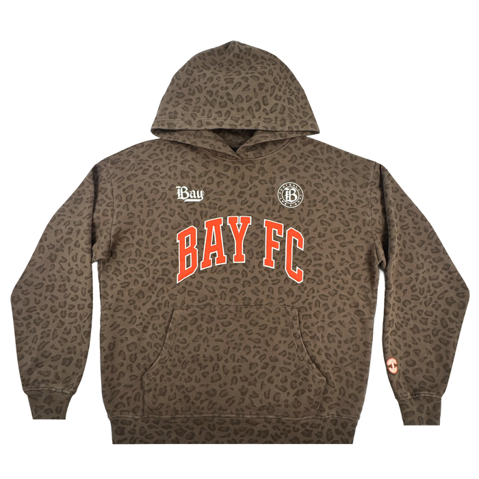 The Bay FC Leopard Hoodie features a brown pullover design with an all-over Leopard Camo print, bold red and white "BAY FC" across the chest, two white logos above, and a small orange symbol on the left sleeve near the wrist.