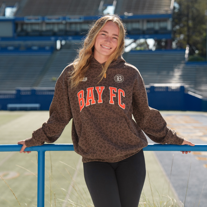 A young woman with long, light brown hair smiles as she leans against a blue railing in an empty stadium, wearing the Bay FC Leopard Hoodie with bold red "BAY FC" lettering and black leggings. Sunlight brightens the scene.