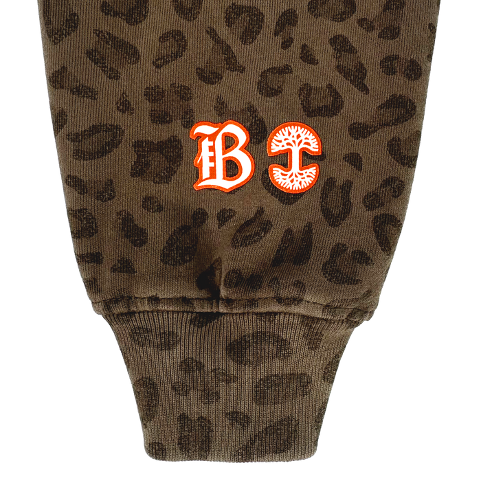 A close-up of the Bay FC Leopard Hoodie shows brown Leopard Camo fabric and a ribbed cuff. Near the cuff are two embroidered designs: a red gothic "B" with white trim, and a red "C" with a stylized mushroom. Brand: Bay FC.