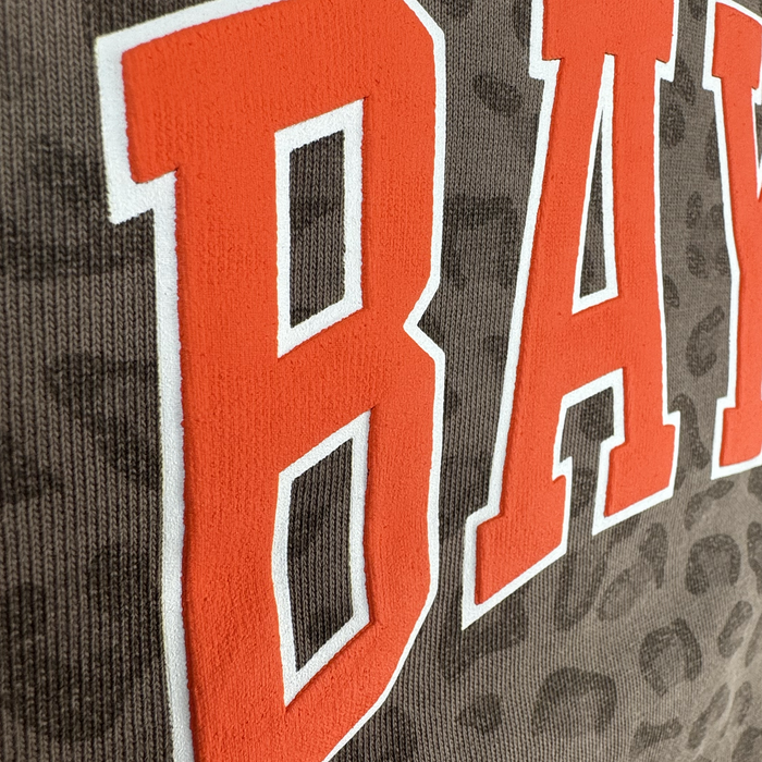 Close-up of the Bay FC Leopard Hoodie shows textured gray fabric with a subtle leopard camo print and bold red-orange “BAN” lettering with white outlines. Visible stitching and ribbed details highlight Bay FC’s signature style.