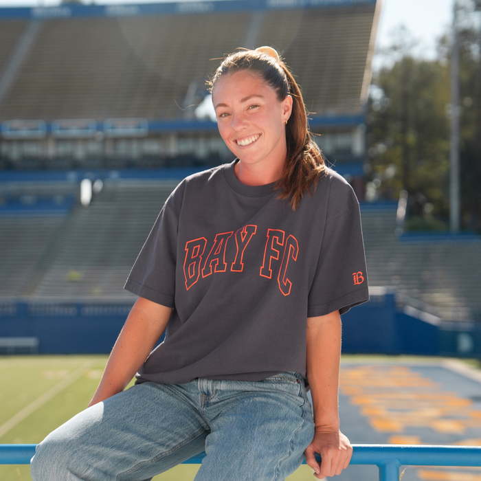 A smiling woman with light skin and brown hair pulled back sits on a blue railing at a stadium, wearing the Bay FC Varsity Tee by Bay FC in dark gray heavy-weight cotton and light blue jeans. Empty seats and a green field shine behind her.