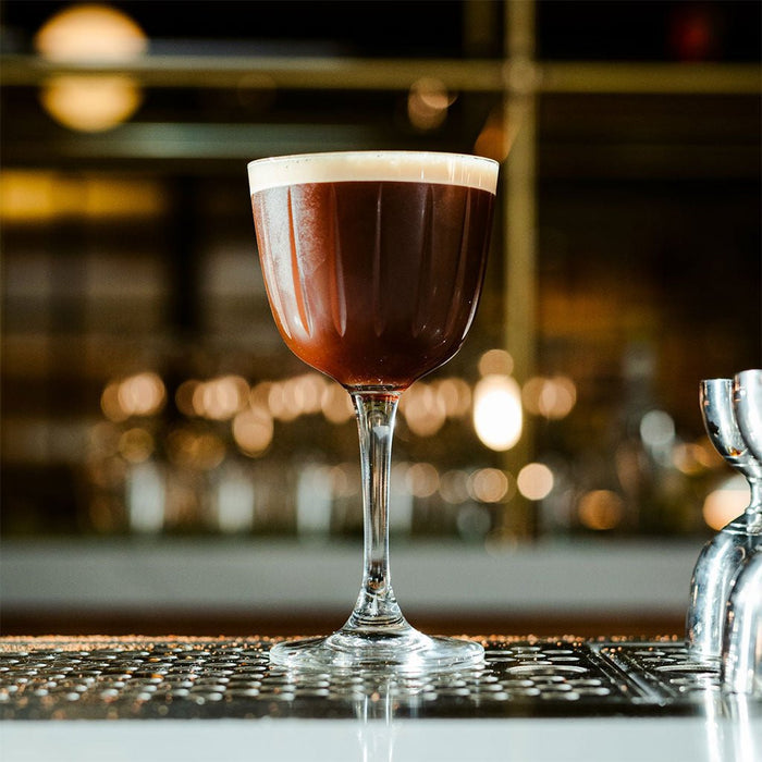 Dark brown cocktail with a white foam top in a glass on a bar counter.