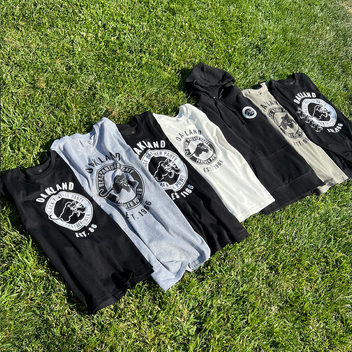 Seven shirts and hoodies with the "Oakland Fight Racism" logo are arranged on green grass, embodying Social Consciousness Apparel. Featuring the "Black Panther Power Tee" by Oaklandish, these garments come in black, gray, and white and display a bear alongside the text "OAKLAND FIGHT RACISM" and "EST. 1998." The shirts are fanned out from left to right.