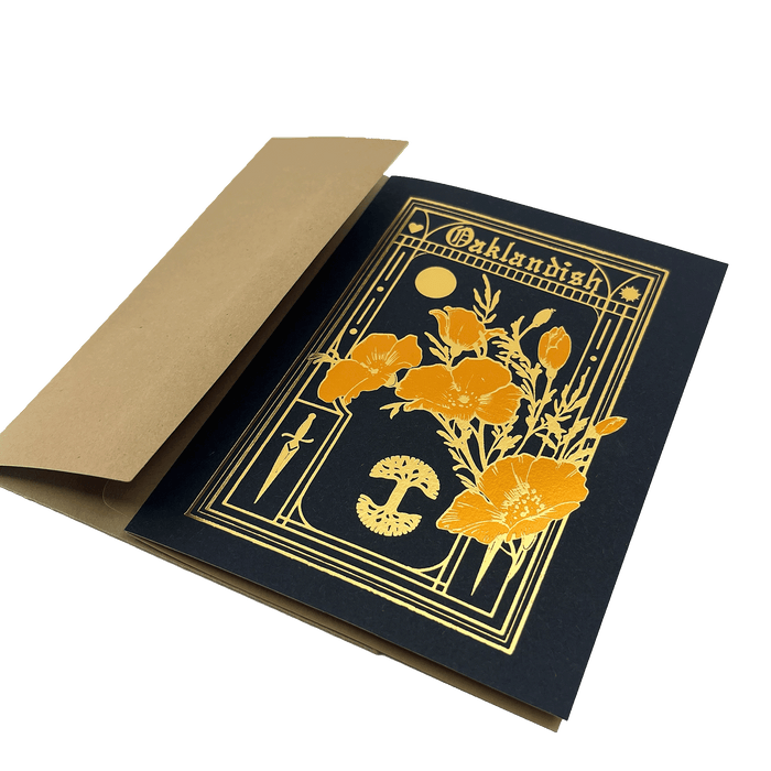 A rectangular Blossom Card by Never Elsewhere features a black background adorned with a gold foil floral design showcasing large poppy flowers and a mushroom. "Oaklandish" is printed in letterpress above the design. The card is partially inserted into a slightly open tan envelope made of recycled kraft paper.