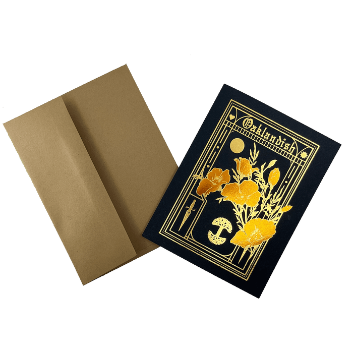 An elegant Blossom Card by Never Elsewhere, featuring a sophisticated black design with a gold-foil arrangement of flowers, mushrooms, a book, and a moon within a rectangular border. The word "Outlandish" is displayed at the top. This charming greeting card comes with a plain brown envelope made from recycled kraft paper.