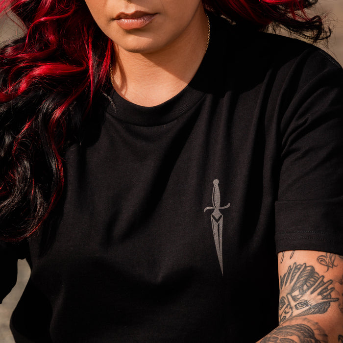 A person with long, dark hair featuring red highlights is wearing the Blossom Tee by Oaklandish—a men's cotton t-shirt adorned with a small sword graphic on the left side of the chest. Their left arm is decorated with various tattoos, including floral motifs and other intricate patterns. The person is sitting outdoors.