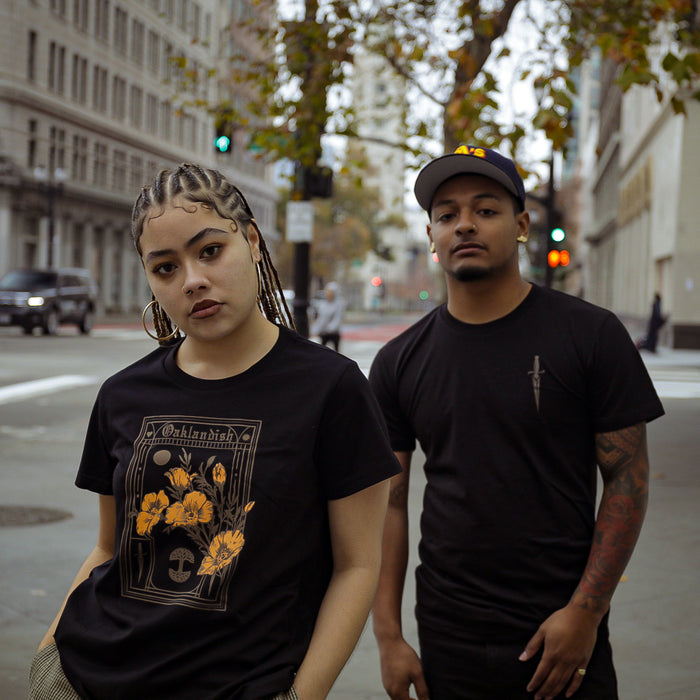 Two individuals stand in the foreground of an urban street scene. The person on the left has braided hair and wears an Oaklandish Blossom Tee, showcasing California poppies, while the person on the right wears a black shirt and a cap. They are on an empty city street with buildings and trees in the background.