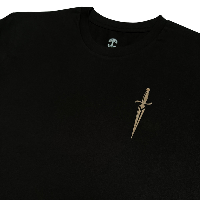 A close-up of the Oaklandish Blossom Tee, a black men's cotton T-shirt featuring a small, intricately designed gold dagger emblem on the upper left chest. The dagger has a sharp, pointed blade and an elegant hilt with stylized details. The plain white background makes the T-shirt and dagger design stand out beautifully.