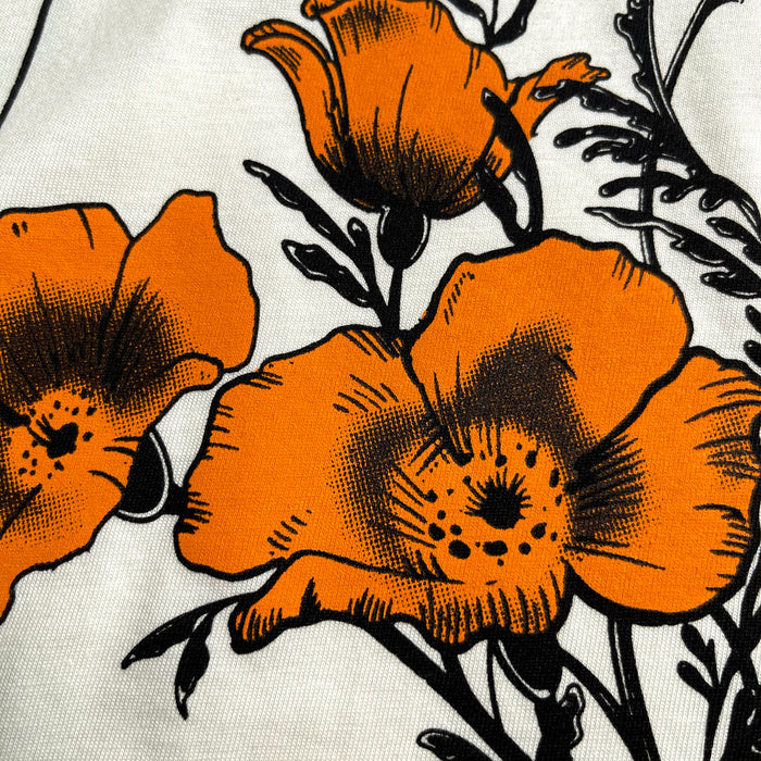 A close-up image of the Oaklandish Blossom Tee, made from 100% cotton and featuring a classic fit, showcases a print adorned with large orange poppy flowers and black outlines on a white background. The vibrant design captures the intricate details of the petals and leaves, creating a striking contrast between the bold colors and the negative space.