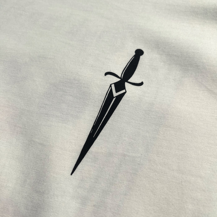 The Blossom Tee by Oaklandish features a minimalist black dagger design centered on a plain white, 100% cotton background. The dagger showcases a straight blade with a geometric pattern near the hilt and a simple crossguard, highlighting the clean, bold contrast on this classic fit t-shirt.