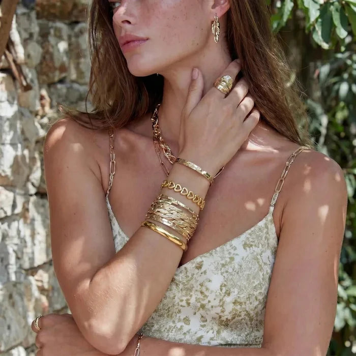 Bold Hinge Bangle in polished gold-tone metal, a slim smooth band worn with layered gold bracelets and rings.