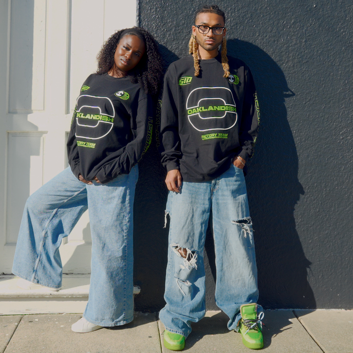 Two people pose by a dark wall, both wearing oversized light blue jeans and black Oaklandish Brrraaap Moto LS Tees. The person on the right pairs theirs with green sneakers and sunglasses for a cool, effortless look.