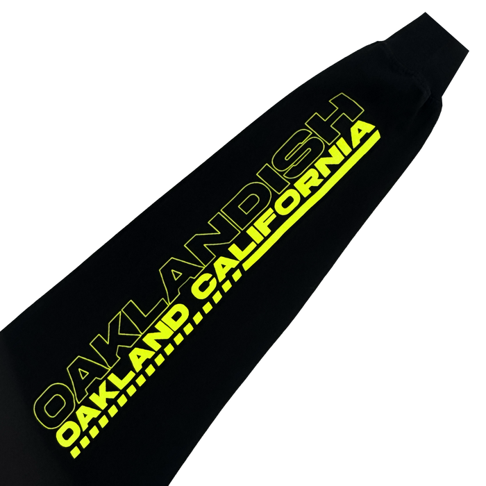 The Brrraaap Moto LS Tee by Oaklandish is a black men’s long sleeve shirt featuring bold, bright yellow “OAKLANDISH” text above smaller “OAKLAND CALIFORNIA” lettering with diagonal lines, shown on a plain white background.