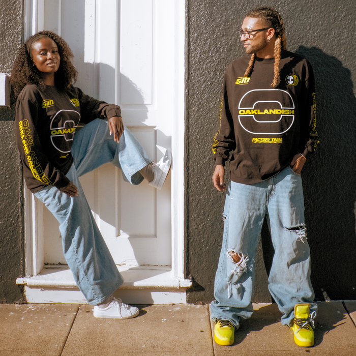 Two people stand by a dark wall near a white door, both in black t-shirts. One wears the Oaklandish Brrraaap Moto LS Tee with baggy light blue jeans and bright yellow sneakers; the other leans on the step, smiling.