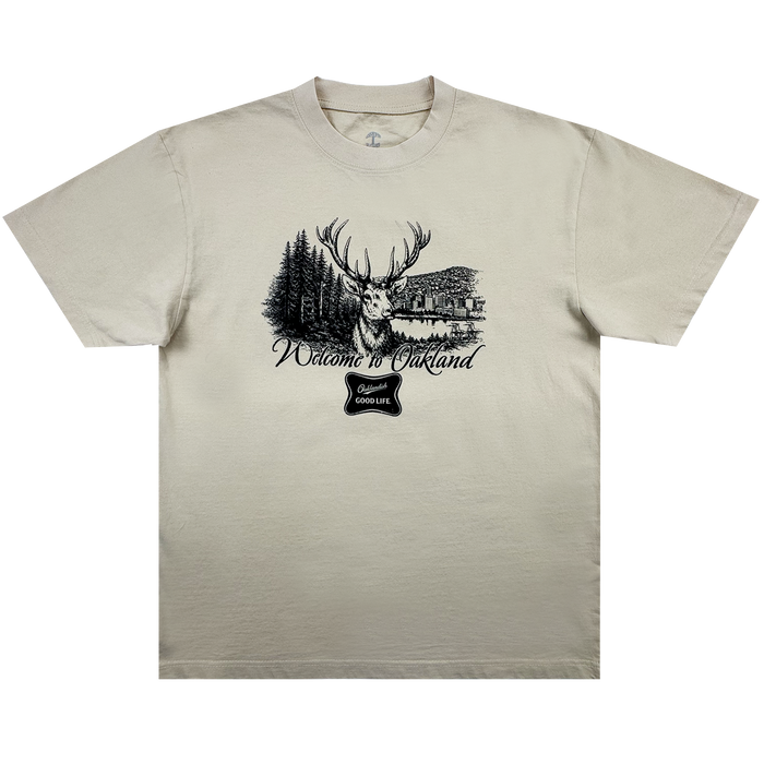 The Buck Tee by Oaklandish is a beige men’s t-shirt with a black stag, pine trees, and lake cabin graphic. Made from heavy-weight cotton, it features "Welcome to Oakland" in cursive and a “good life” badge below.