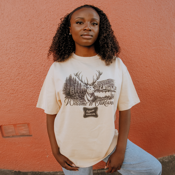 A woman with curly hair stands against a textured orange wall, wearing the Oaklandish Buck Tee, a classic fit cream t-shirt made from heavy-weight cotton. It features a deer, trees, building, and "Welcome to Oakland" and "Good Life" prints.