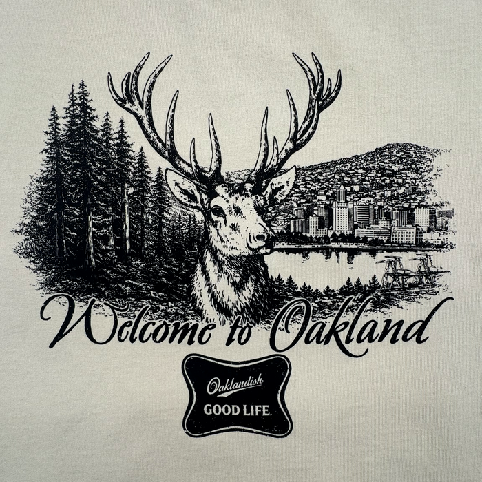 The Oaklandish Buck Tee features a black and white deer with large antlers before pine trees and a cityscape. "Welcome to Oakland" text appears above, with an "Oaklandish GOOD LIFE" badge below. Part of the men's collection.