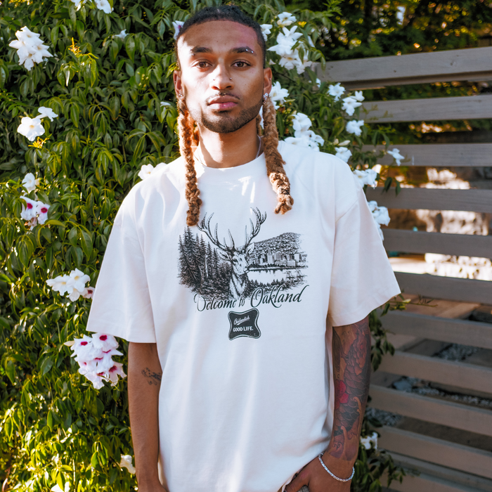 A man with long, braided hair stands in sunlight, wearing the Oaklandish Buck Tee—a classic fit shirt crafted from heavy-weight cotton featuring a graphic of a deer, trees, a building, and "Welcome to Oakland." White flowers line the wooden fence behind him.