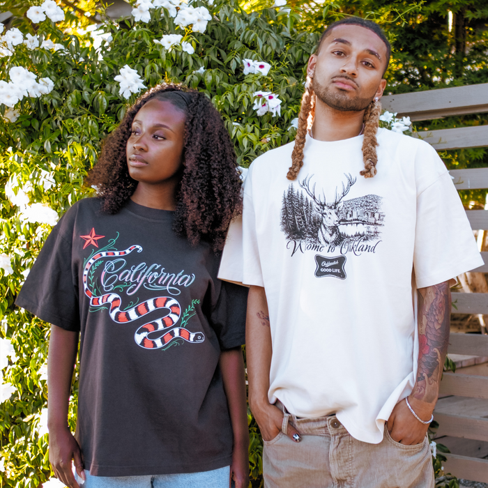 Two people stand outdoors before greenery. The left wears a black "California" snake tee, while the right sports Oaklandish's Buck Tee, a heavy-weight cotton shirt with an elk and forest illustration.