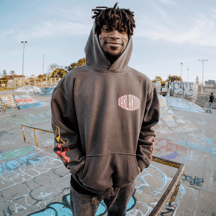 A smiling man with medium-dark skin and short dreadlocks stands at a graffiti-covered skatepark, wearing an oversized, drop shoulder Chopper Heavy Hoodie from Other. His hands are tucked into the pockets of the pullover, and ramps, rails, and trees appear in the background beneath a partly cloudy sky.