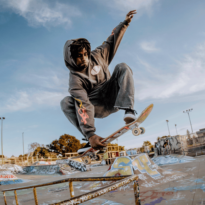 Wearing the Other Chopper Heavy Hoodie in gray and jeans, a skateboarder executes a high-flying trick over a rail at a graffiti-adorned skate park, caught mid-air against a blue sky dotted with clouds. Trees and light poles can be seen in the background.