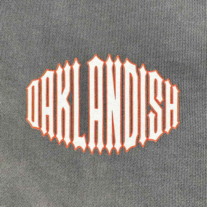 The image features the word “OAKLANDISH” in large, stylized white letters with an orange outline, set against the gray fabric of the Chopper Heavy Hoodie by Other. The elongated, pointed lettering creates a bold and eye-catching look that perfectly complements the oversized drop shoulder design of this pullover hoodie.