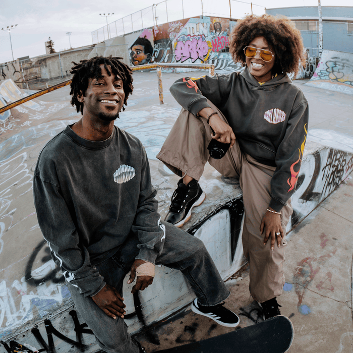 Two people at a graffiti-covered skatepark smile at the camera. One, dressed in the Other brand Chopper Heavy Hoodie—a cozy, oversized drop shoulder pullover—sits on a ledge holding a skateboard; the other stands with one foot up, wearing sunglasses and holding a camera. Both wear casual streetwear in dark tones and enjoy the urban outdoor setting.