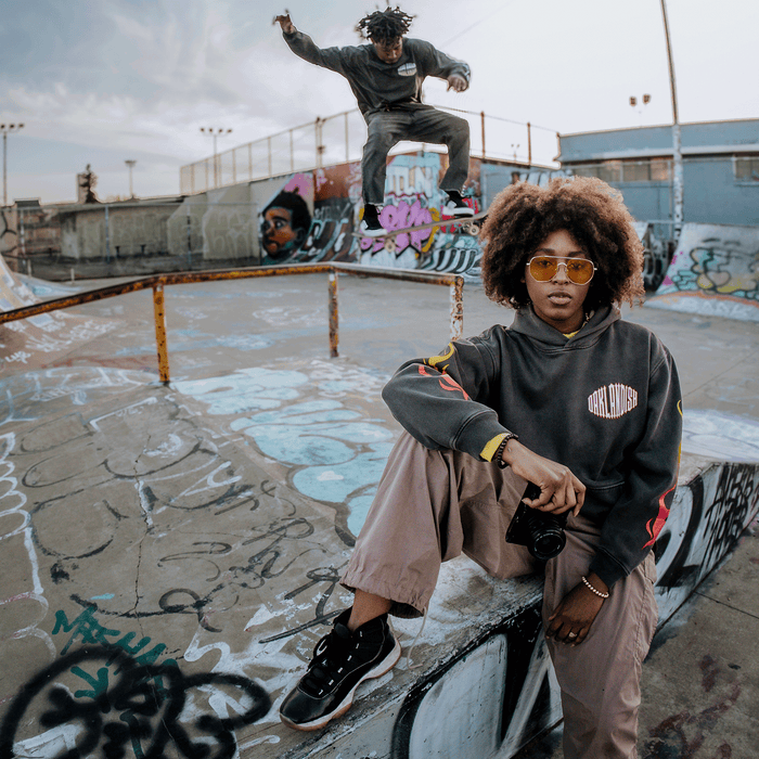 A person with an afro, sunglasses, and a camera, dressed in the Other Chopper Heavy Hoodie with its oversized drop shoulder fit, sits on a graffiti-covered skatepark ramp. Behind them, another skater catches air mid-trick against graffiti walls beneath an overcast urban sky.