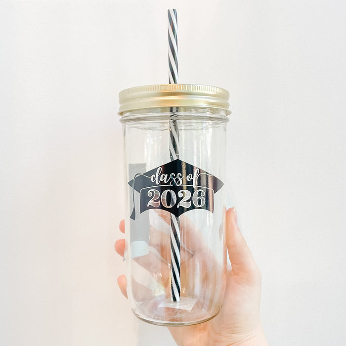 Hand holding a mason jar glass tumbler that has a black grad cap with clear text in the middle that reads class of 2026. Black and white striped straw