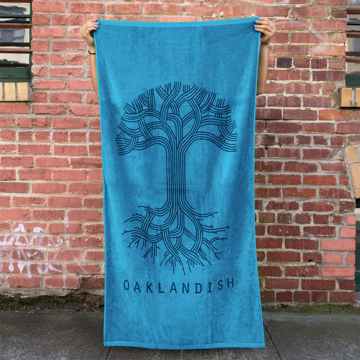 A person is holding up an oversized Classic Logo Beach Towel by Oaklandish, made of cotton terry velour in turquoise. The towel features a stylish dark tone-on-tone print of a stylized tree with intricate branches and roots, with the word "OAKLANDISH" printed at the bottom. The background showcases a rustic brick wall adorned with windows and visible graffiti.