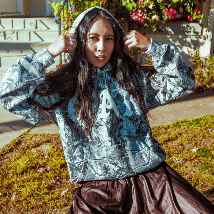 A woman with long dark hair sits outside on a sunny day, pulling up the hood of her Oaklandish Cork Hoodie, which features a grey wood camo pattern and bold white letters. She pairs it with a brown skirt and multiple rings, surrounded by flowers and greenery.
