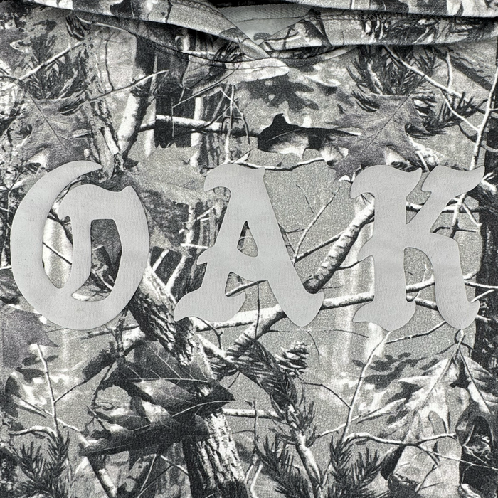 The Oaklandish Cork Hoodie features a grey wood camo design with forest-inspired branches, leaves, and pine cones. Bold "OAK" lettering in light gray makes this men’s outerwear stylish and unique.