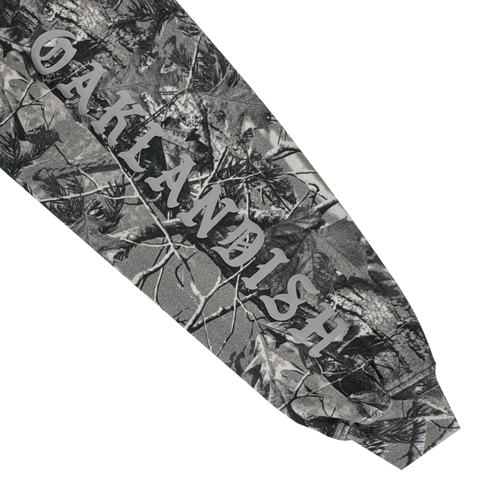 A close-up of the Cork Hoodie by Oaklandish shows a grey wood camo forest pattern. "OAKLANDISH" is printed in large, white serif letters diagonally across the men's outerwear sleeve, surrounded by tree branches and leaves.