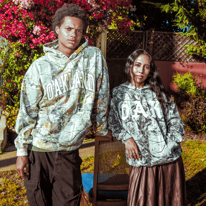 Two people stand outdoors in sunlit gardens, both wearing Oaklandish Cork Hoodies. The left wears cargo pants; the right, a long skirt. They lean on a chair amid blooming flowers and a wooden fence, highlighting these stylish hoodies.