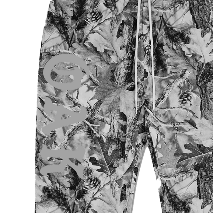 A close-up of the Cork Pant by Oaklandish in 100% cotton, showcasing a grey wood camo pattern with leaves, branches, and pine cones. "OAK" is printed vertically on the left leg and white drawstrings detail the waistband.