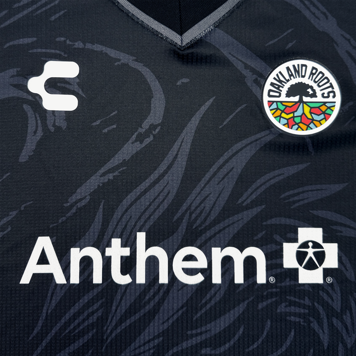 Close-up of the Custom Unisex Oak-ness Awakens Kit by Oakland Roots SC: a dark sports jersey with Oakland folklore-inspired circular logo—a white stylized tree and colorful roots—plus “Anthem,” a medical cross logo, subtle patterns, and a white “C” logo.