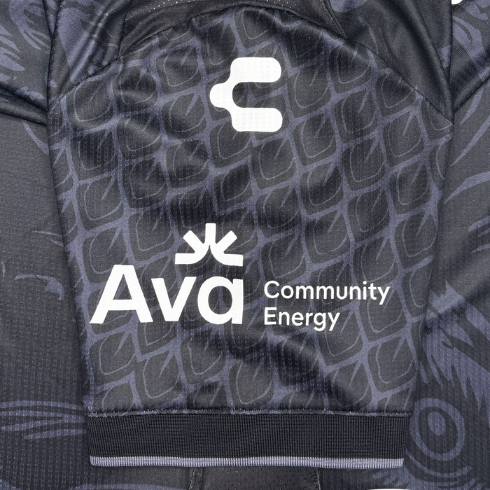 A close-up of the Oakland Roots SC Custom Unisex Oak-ness Awakens Kit, a black jersey with a subtle geometric pattern. The sleeve features white “C” and “Ava Community Energy” logos, with Ava’s sunburst celebrating Oakland folklore and Lake Merritt.
