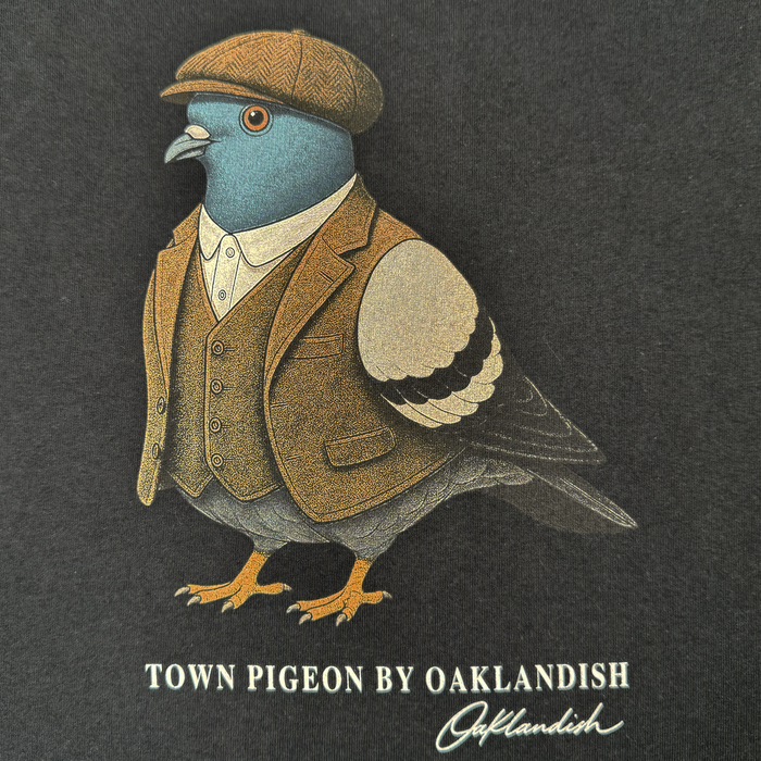A pigeon sporting a brown tweed suit, vest, and flat cap stands upright against a dark background, capturing the timeless style of the men’s collection. Below it reads “Dapper Tee by Oaklandish” with the Oaklandish logo signature underneath—ideal for a classic fit t-shirt in heavyweight cotton.