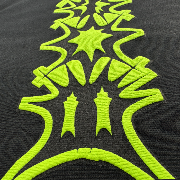 Close-up of neon green embroidery on black textured cotton fleece from the Distortion Hoodie by Other. The symmetrical, geometric design—featuring zigzags, stars, curves, and bold lines—stands out across the oversized drop-shoulder silhouette, creating a striking modern effect with raised stitching.