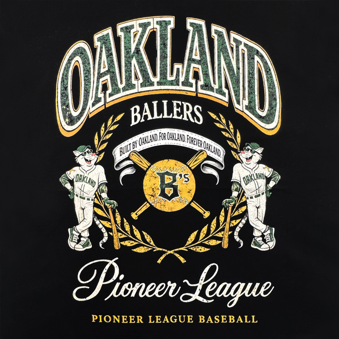 A graphic on the Oakland Ballers Double Tee features two smiling baseball mascots in Oakland uniforms, a golden bat-and-ball crest, and green/yellow accents, with "Oakland Ballers, Pioneer League Baseball" text celebrating the team.