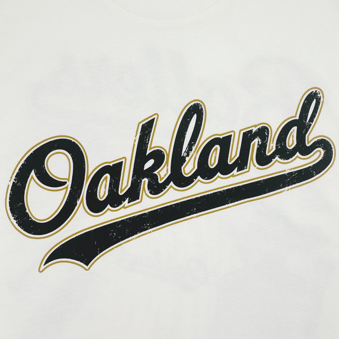 The Dugout Tee by Oakland Ballers features “Oakland” in bold black cursive with a gold outline and distressed finish on white, inspired by classic Oakland baseball team logos.