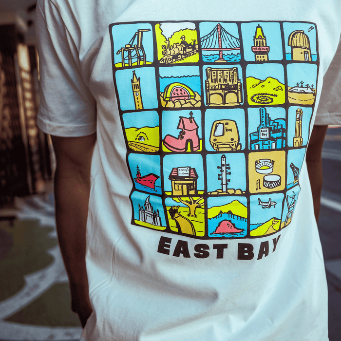 Someone wears the Oaklandish East Bay Landmarks Tee, a white shirt showcasing colorful cartoon squares of Bay Area bridges, buildings, and whimsical scenes on blue and yellow backgrounds.
