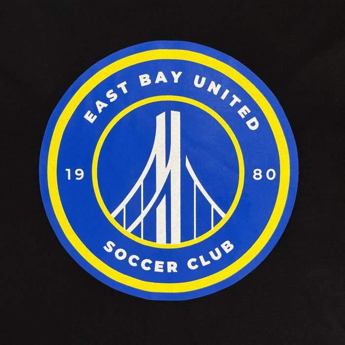 The EBU Women's Tee by Supported Brands showcases a circular logo featuring "East Bay United Soccer Club" in white text on a blue background. The year "1980" is displayed with "19" on the left and "80" on the right. At the center, a stylized white suspension bridge is set against a blue backdrop with a yellow outer ring, representing this vibrant youth soccer club.
