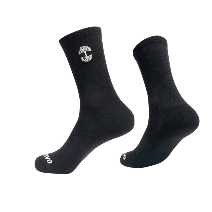 A pair of black crew socks, with one sock slightly elevated and angled to the side. The Embroidered Logo Socks feature a small, white, vertical emblem resembling a tree on the upper part and white text "OAKPO" near the toes. These Oaklandish socks come in adult sizes, boasting a ribbed texture on top and smooth finish below.
