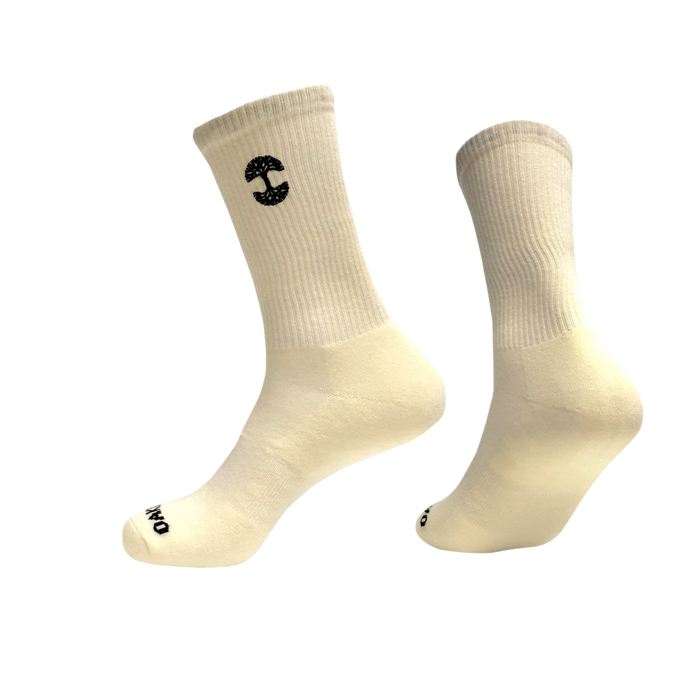 Two off-white cotton crew socks with black logos are suspended mid-air against a white background. The Oaklandish Embroidered Logo Socks are ribbed on the calf area, with one displaying an embroidered stylized "C" near the top and the other sporting a small circular logo on the toe. The overall design is minimalistic and clean.